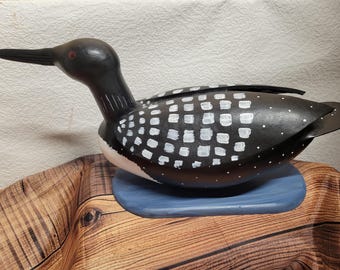 May include: A hand-painted wooden loon sculpture. The bird has a black head and back with white square markings. It has a long black beak and a red eye. The loon is mounted on a blue oval base.