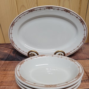 Może przedstawiać: Biały owalny półmisek i trzy małe białe miseczki. Każdy element ma brązową kwiatową obwódkę. Półmisek jest podparty złotym metalowym stojakiem. Naczynia są ułożone na drewnianej powierzchni.