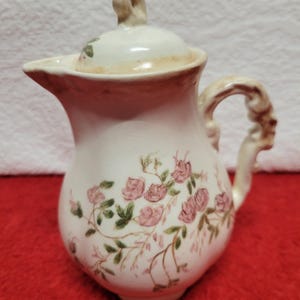Op de afbeelding: Crèmekleurige keramische creamer met deksel, versierd met roze rozen en groene bladeren. De creamer heeft een gebogen handvat en een schenktuit. Een decoratief item voor het serveren.