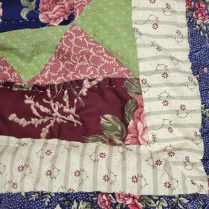 May include: Close-up of a patchwork quilt with floral and geometric patterns. The quilt features a variety of fabrics in shades of blue, green, burgundy, and cream. The design includes large floral prints, small floral prints, and striped borders. The quilt is a mix of textures and colors.