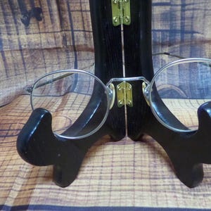 Puede incluir: Gafas redondas con montura plateada expuestas en un soporte de madera negro. Las gafas tienen un diseño clásico y minimalista con monturas finas y lentes transparentes. El soporte es de madera oscura y brillante con bisagras de latón, sobre un fondo de vetas de madera.