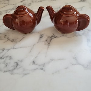 May include: Two brown ceramic teapot-shaped salt and pepper shakers. Each shaker has a spout, handle, and a small opening on top for dispensing. The shakers are on a white and gray marble surface.