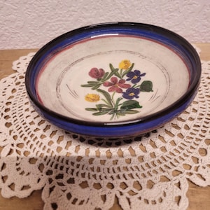 May include: A ceramic bowl with a floral design. The bowl has a white interior with a painted bouquet of purple, red, and yellow flowers. The rim is blue and black. The bowl is sitting on a white lace doily.