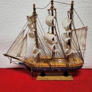 May include: A detailed wooden model of a sailing ship. The ship features white sails, brown masts, and a blue hull. The model includes intricate rigging and an anchor, resting on a wooden base. The ship is a decorative nautical item.