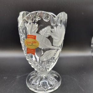 May include: A clear crystal vase with a scalloped rim and a pedestal base. The vase is adorned with leaf and floral designs. A red and gold label on the front reads "Anna Hütte Bleikristall" and "24% PbO".