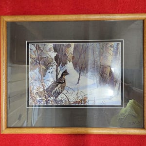 May include: Framed print of a bird in a snowy forest scene. The artwork features a brown and white bird perched on a branch, with a backdrop of snow-covered trees and a small building. The frame is made of light brown wood.