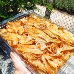 May include: A rectangular clear plastic container filled with thin, translucent, golden-brown dried fish strips seasoned with small red chili flakes and yellow spice specks. The dried seafood snack is stacked in layers within the container.