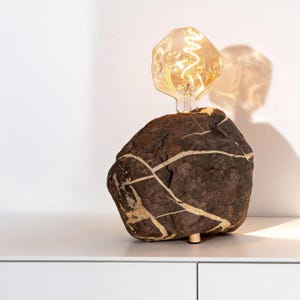 Könnte beinhalten: Eine einzigartige Tischlampe mit einem großen, dunkelbraunen Felsen mit weißen Adern. Die Lampe hat eine dekorative, geometrische Glühbirne. Die Lampe steht auf einer weißen Oberfläche. Der Felsen ist etwa 25 cm hoch.