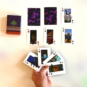 May include: A deck of playing cards with a Minecraft theme. The cards feature various characters and scenes from the game. The card backs have a purple pixelated design. The deck box has a pixelated tree design.