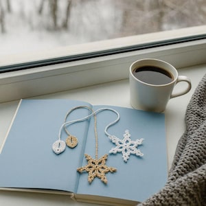 May include: A cozy scene featuring a light blue book with three crochet snowflake bookmarks. A white mug of coffee sits nearby, with a gray knitted blanket and a snowy window in the background. The bookmarks are white, gold, and beige.