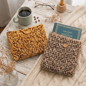 May include: Two rectangular crochet pouches with wooden button closures. One pouch is a solid mustard yellow, and the other is a variegated brown and beige, containing a blue book titled Persuasion by Jane Austen, alongside a coffee mug and glasses.