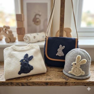 May include: A cream-colored sweater, navy blue crossbody bag, and gray beanie, each adorned with a bunny applique. The sweater and beanie are knit, while the bag has a leather trim. The items are arranged on a wooden surface.