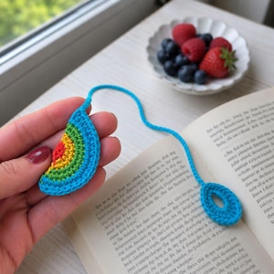 May include: A handmade crochet bookmark in the shape of a teardrop with a rainbow design. The bookmark is held over an open book. The bookmark has a blue string and a blue circular end.