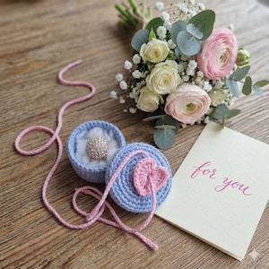 Puede incluir: Una caja de anillos de ganchillo azul claro con un anillo brillante, junto a un ramo de flores y una tarjeta con la frase "for you". La caja de anillos tiene un corazón rosa. Las flores son blancas y rosas.