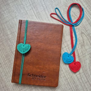 May include: A brown leather notebook with the Schneider Electric logo and a teal crochet heart bookmark. Two additional crochet heart bookmarks, one red and one blue, are also visible.