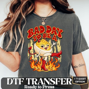 May include: A charcoal gray t-shirt featuring a graphic of a deviled egg character with red horns, a red tail, and red cowboy boots, standing in flames. The text above reads "BAD DAY TO BE A" in a bold, red, wavy font. The shirt has a crew neckline.