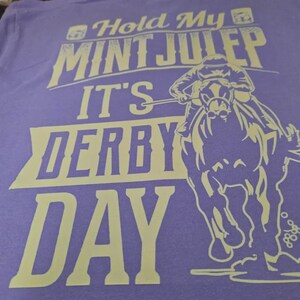 May include: A light purple t-shirt featuring a cream-colored graphic of a jockey riding a horse. The text reads "Hold My MINT JULEP IT'S DERBY DAY" in a bold, stylized font. The design is centered on the fabric, showcasing a horse race theme.