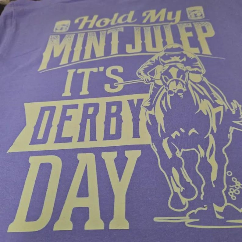 May include: A light purple t-shirt featuring a cream-colored graphic of a jockey riding a horse. The text reads "Hold My MINT JULEP IT'S DERBY DAY" in a bold, stylized font. The design is centered on the fabric, showcasing a horse race theme.