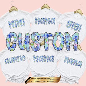 May include: A collection of white t-shirts featuring custom text in a blue floral pattern. The shirts display words including Mimi, Mama, Gigi, Auntie, and Nana. A large, central graphic reads CUSTOM in the same blue floral print. A banner reads NEW DTF TRANSFER or PRINTED T-SHIRT.