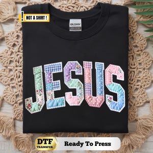 May include: A black sweatshirt featuring the word JESUS in large, patchwork-style letters. Each letter is composed of various pastel-colored fabrics with floral, gingham, and polka dot patterns, outlined with white stitching. Includes a NOT A SHIRT label.