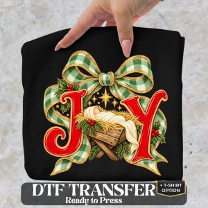 May include: A black fabric transfer featuring the red letters J and Y, with a central graphic of a baby in a manger surrounded by a green and white gingham bow, holly leaves, and berries. The text DTF TRANSFER + T-SHIRT OPTION is below.