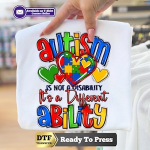 May include: A white t-shirt featuring a colorful graphic with the text autism is not a disability it's a different ability. The design includes a large heart filled with a multi-colored puzzle pattern, surrounded by smaller green and yellow hearts.