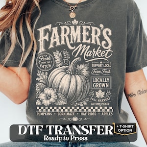May include: A charcoal gray t-shirt featuring a white graphic print of a pumpkin and the text Farmer's Market, Fresh From The Patch, Support Local, Farm Fresh, Locally Grown, Fall Harvest, Autumn Picked, Pumpkins, Corn Maze, Hay Rides, Apples.