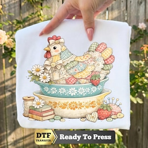 May include: A white fabric transfer featuring a patchwork chicken with a bow sitting in a teal bowl, stacked on a yellow bowl. The design includes daisies, a stack of books, a spool of thread, strawberries, and buttons with the text DTF Ready To Press.