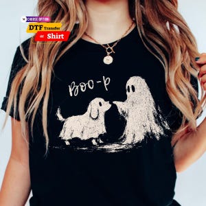 May include: A black t-shirt featuring a white graphic print of a ghost and a dog, with the text "Boo-p" written above them. The design has a distressed, textured appearance. The shirt is shown with a gold chain necklace.