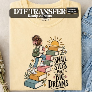 May include: A pale yellow t-shirt featuring a graphic of a person with a backpack climbing a staircase made of colorful books. The text reads DTF TRANSFER Ready to Press + T-SHIRT OPTION and SMALL STEPS MAKE BIG DREAMS with sun, flower, and star accents.