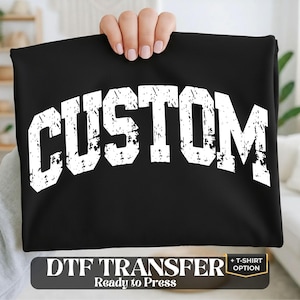 May include: A black fabric sheet featuring the word CUSTOM in large, white, distressed varsity-style lettering. Below the fabric, a black rectangular label displays the white text DTF TRANSFER Ready to Press + T-SHIRT OPTION.