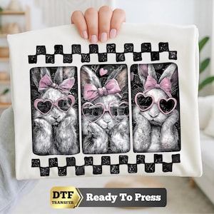 May include: A white fabric sheet featuring a graphic print of three rabbits in separate panels. Each rabbit has a pink bow and pink heart-shaped sunglasses. The design is framed by a black and white checkered border. Text at the bottom reads DTF TRANSFER Ready To Press.