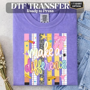 May include: A purple t-shirt featuring a graphic print with vertical stripes, a pink bow, a yellow pencil, a yellow ruler, and a yellow school bus. The text reads make a difference in white and colorful script lettering on a school-themed background.