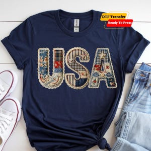 May include: A navy blue t-shirt featuring the letters USA in a patchwork design. The letters are filled with various floral, plaid, and gingham fabric patterns in red, white, and blue, outlined with cream lace trim and small pearl accents.