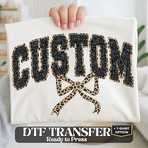 May include: A white t-shirt featuring the word CUSTOM in black glittery varsity-style letters with a leopard print outline. Below the text is a large leopard print bow graphic. The image includes the text DTF TRANSFER + T-SHIRT OPTION.
