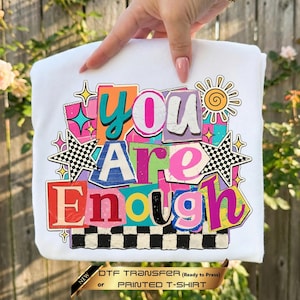 May include: A white t-shirt featuring a colorful graphic with the text "You Are Enough" in various fonts and bright colors. The design includes checkered stars, a sun icon, and geometric shapes in pink, orange, blue, and green on a white background.