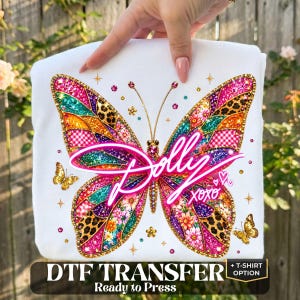 May include: A white t-shirt featuring a large, colorful butterfly graphic with intricate patterns including leopard print, floral designs, and glittery textures. The word Dolly is written in a bright pink script across the center, with XOXO below it.