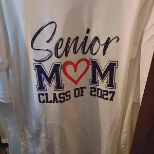 May include: A white t-shirt featuring dark blue text that reads Senior, followed by the letters M and M separated by a red heart graphic, and the text CLASS OF 2027 below. The design is printed in a combination of script and block lettering.