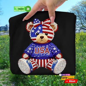 A sparkling faux rhinestone teddy bear design with American flag patterns on its face and bow, wearing a blue USA sweater and red-white striped pants. This high-quality patriotic DTF transfer and custom shirt option is perfect for the USA 250th anniversary celebrations, 4th of July parades, and Independence Day events. The 250 years of America commemorative graphic features a glittery diamond effect for a luxury look on black t-shirts and hoodies.