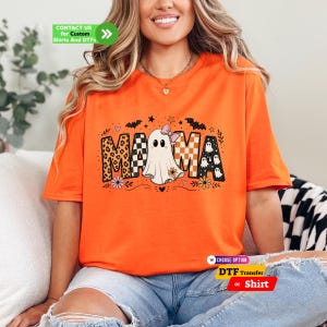 May include: An orange t-shirt featuring a graphic print of the word MAMA, where the letter A is replaced by a white ghost wearing a pink bow. The letters include leopard print, checkered patterns, and small ghost motifs with bats and flowers.