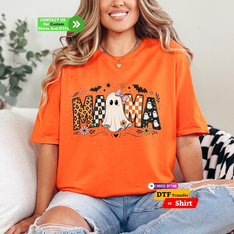 May include: An orange t-shirt featuring a graphic print of the word MAMA, where the letter A is replaced by a white ghost wearing a pink bow. The letters include leopard print, checkered patterns, and small ghost motifs with bats and flowers.