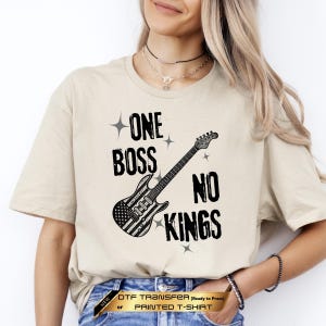 May include: A beige short-sleeved t-shirt featuring a black graphic print of an electric guitar with an American flag pattern on the body. The text reads ONE BOSS NO KINGS, accompanied by small four-pointed star icons surrounding the central design.