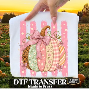 May include: A white t-shirt featuring a graphic of a pumpkin with a pink glitter bow. The pumpkin has segments with leopard print, floral patterns, stars, and gingham. It is set against vertical pink brushstrokes and small gold sparkles.