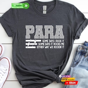 May include: A dark heather gray short-sleeve t-shirt featuring the white text PARA in a checkered pattern above the text SOME DAYS I ROCK IT SOME DAYS IT ROCKS ME EITHER WAY WE ROCKIN IT with three horizontal lines and lightning bolt icons.