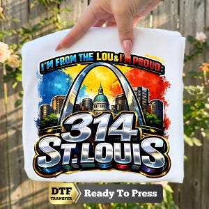 May include: A white t-shirt featuring a graphic print of the St. Louis Gateway Arch and city skyline. The text reads I'M FROM THE LOU & I'M PROUD, 314, and ST. LOUIS. The design includes vibrant blue, yellow, and red watercolor-style splashes.