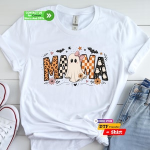 May include: A white short-sleeve t-shirt featuring a graphic that spells MAMA, where the letter A is replaced by a ghost wearing a pink bow. The letters feature leopard print, black and white checkers, and orange patterns with small ghosts, bats, and flowers.