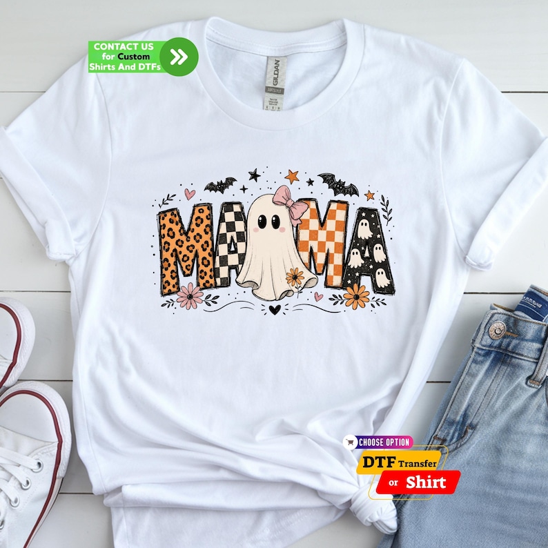 May include: A white short-sleeve t-shirt featuring a graphic that spells MAMA, where the letter A is replaced by a ghost wearing a pink bow. The letters feature leopard print, black and white checkers, and orange patterns with small ghosts, bats, and flowers.