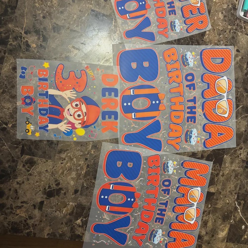 May include: Four transparent sheets featuring colorful birthday-themed graphics in orange and blue. Designs include text such as DADA OF THE BIRTHDAY BOY, MAMA OF THE BIRTHDAY BOY, and DEREK 3 BIRTHDAY BOY, alongside illustrations of cakes, sunglasses, and a cartoon character.