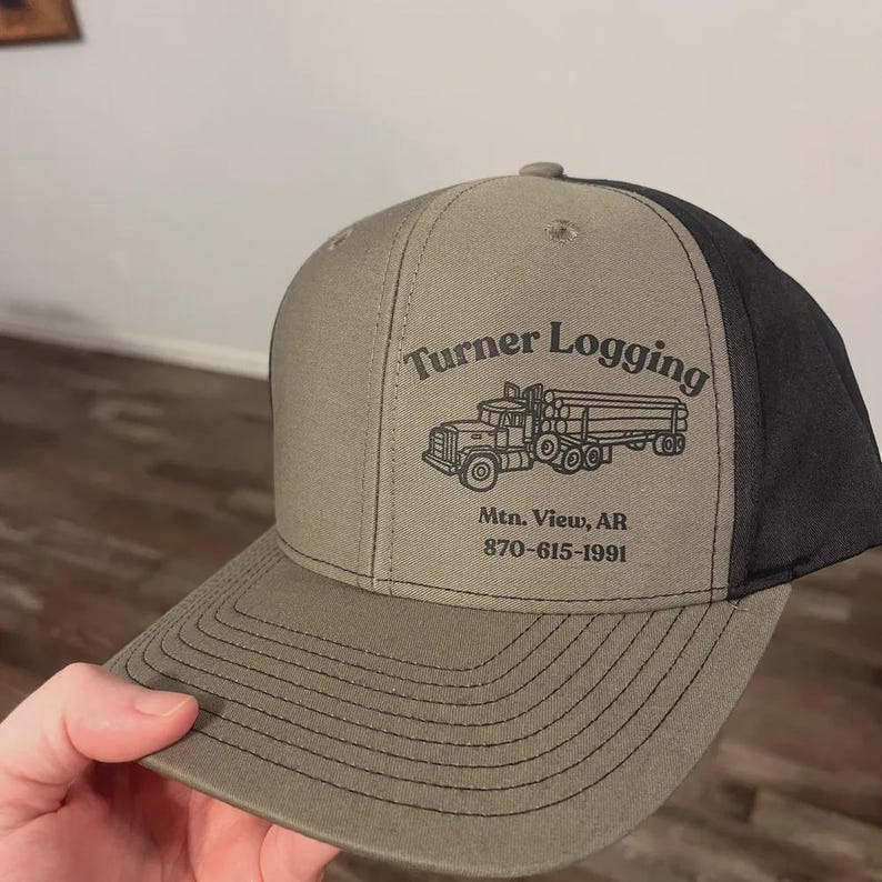 May include: A khaki and black baseball cap featuring a black graphic of a logging truck and the text Turner Logging, Mtn. View, AR, and 870-615-1991 printed on the front panel. The cap has a structured crown, a flat brim with stitching, and black mesh.