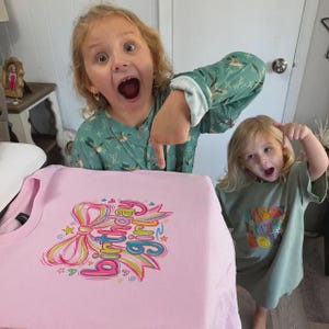 May include: A light pink t-shirt featuring a colorful graphic print of a large bow and the text "birthday girl" in a stylized font. The design includes small star and heart accents in shades of yellow, blue, green, and bright pink.
